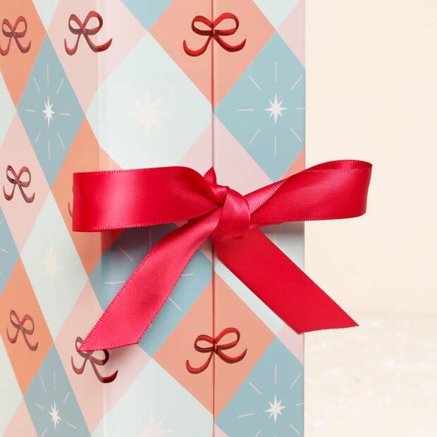 A close-up of a gift box wrapped in pastel argyle holiday paper with red bow motifs and tied with a large red satin ribbon bow.
