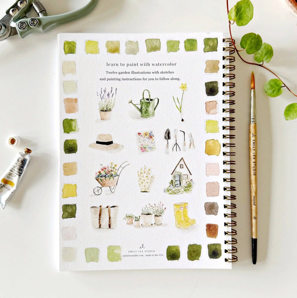 Notebook with watercolor illustrations of garden-themed designs on a white surface with art supplies.