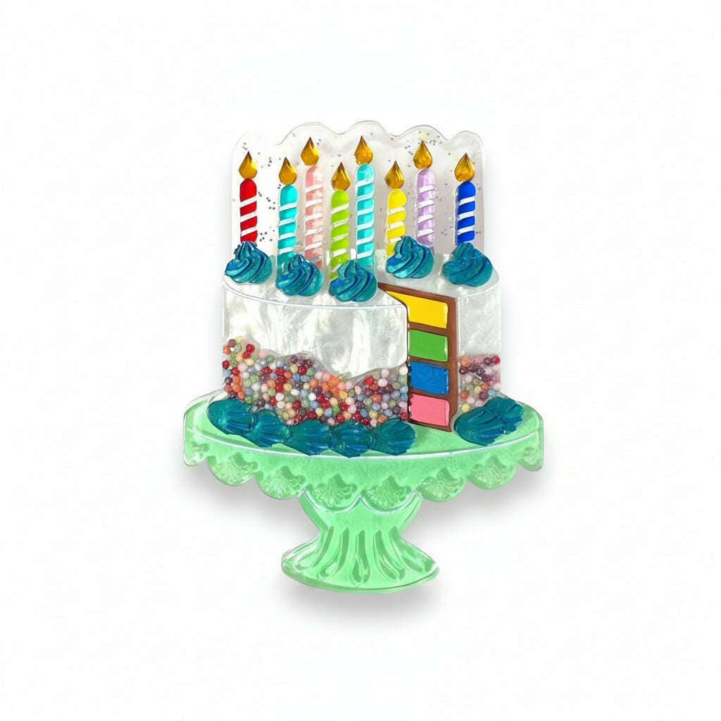A novelty birthday cake-shaped accessory on a mint green pedestal stand, featuring clear acrylic layers with blue frosting swirls, colorful striped candles, and multicolored sprinkles, set against a plain white background.