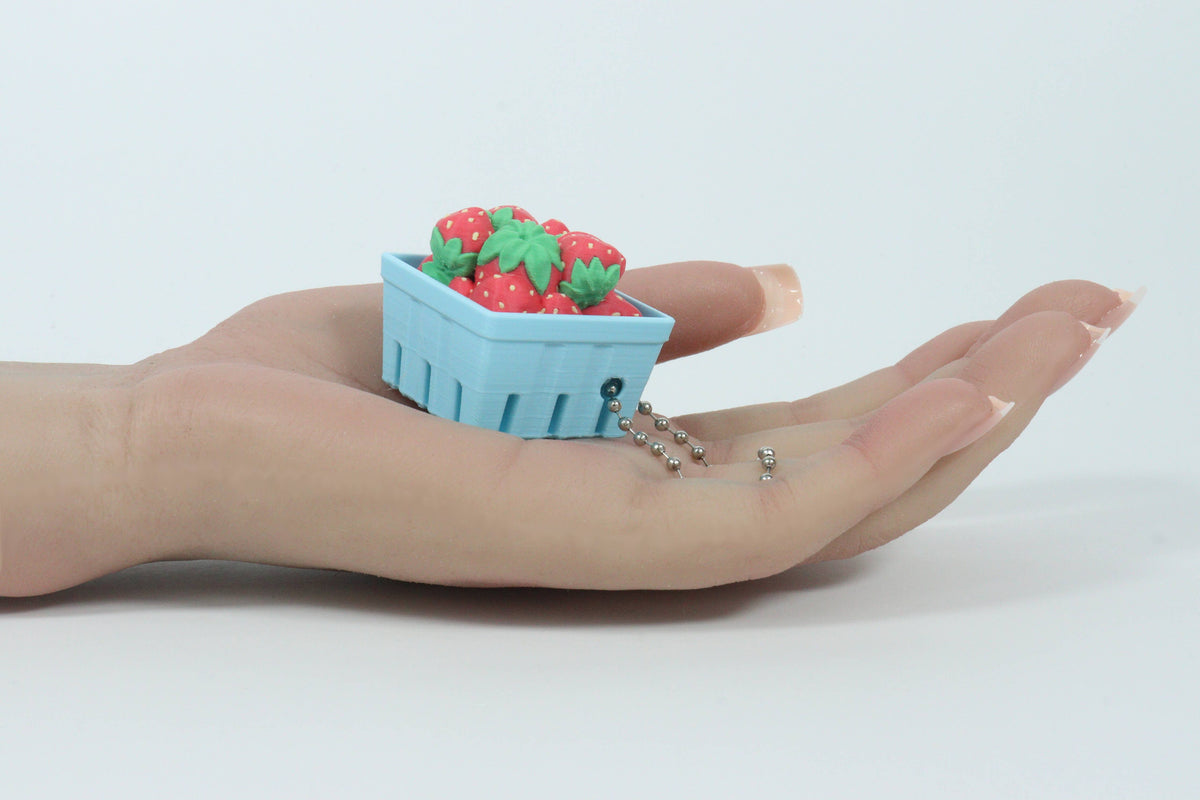 A miniature light blue berry basket 3D-printer charm filled with red strawberries and green leaves, resting on a person's open palm. The charm is attached to a clear strand with small silver beads.