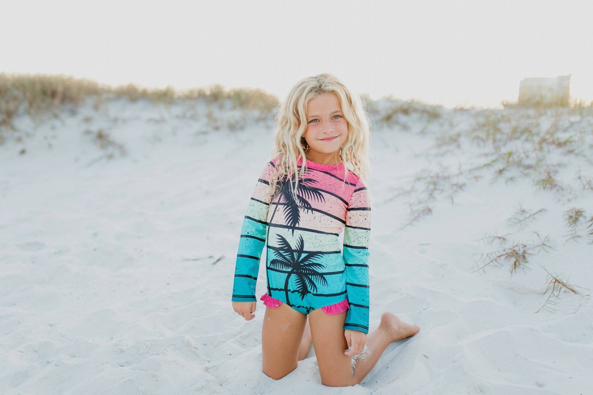 Girl in long-sleeve rash guard swimsuit with palm tree print on beach.
