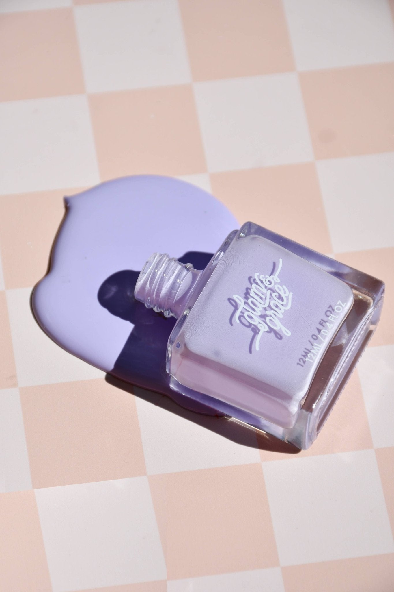 Glam & Grace light purple nail polish bottle with spilled 12ml / 0.4 fl oz polish on a checkered surface.