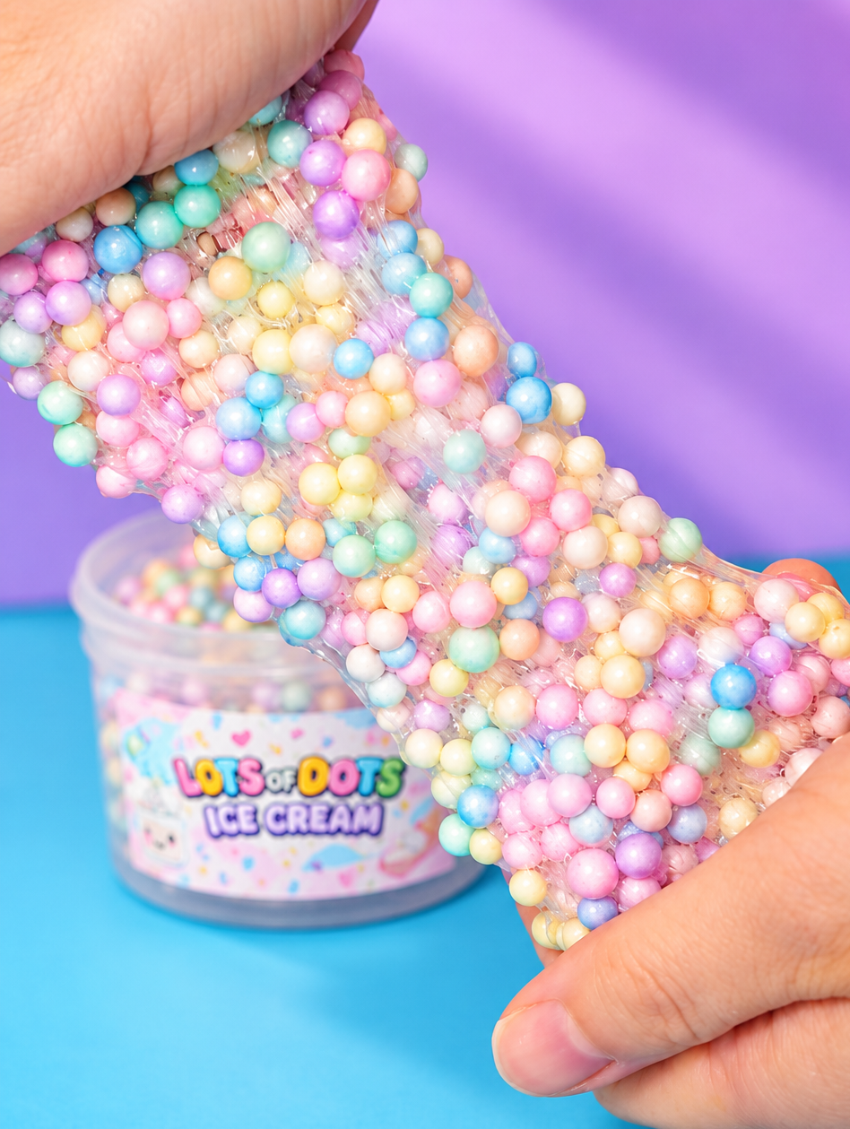 A close-up of hands stretching a translucent slime covered in pastel rainbow foam beads, with a small clear jar labeled 'LOTS OF DOTS ICE CREAM' in the background against a purple and blue backdrop.