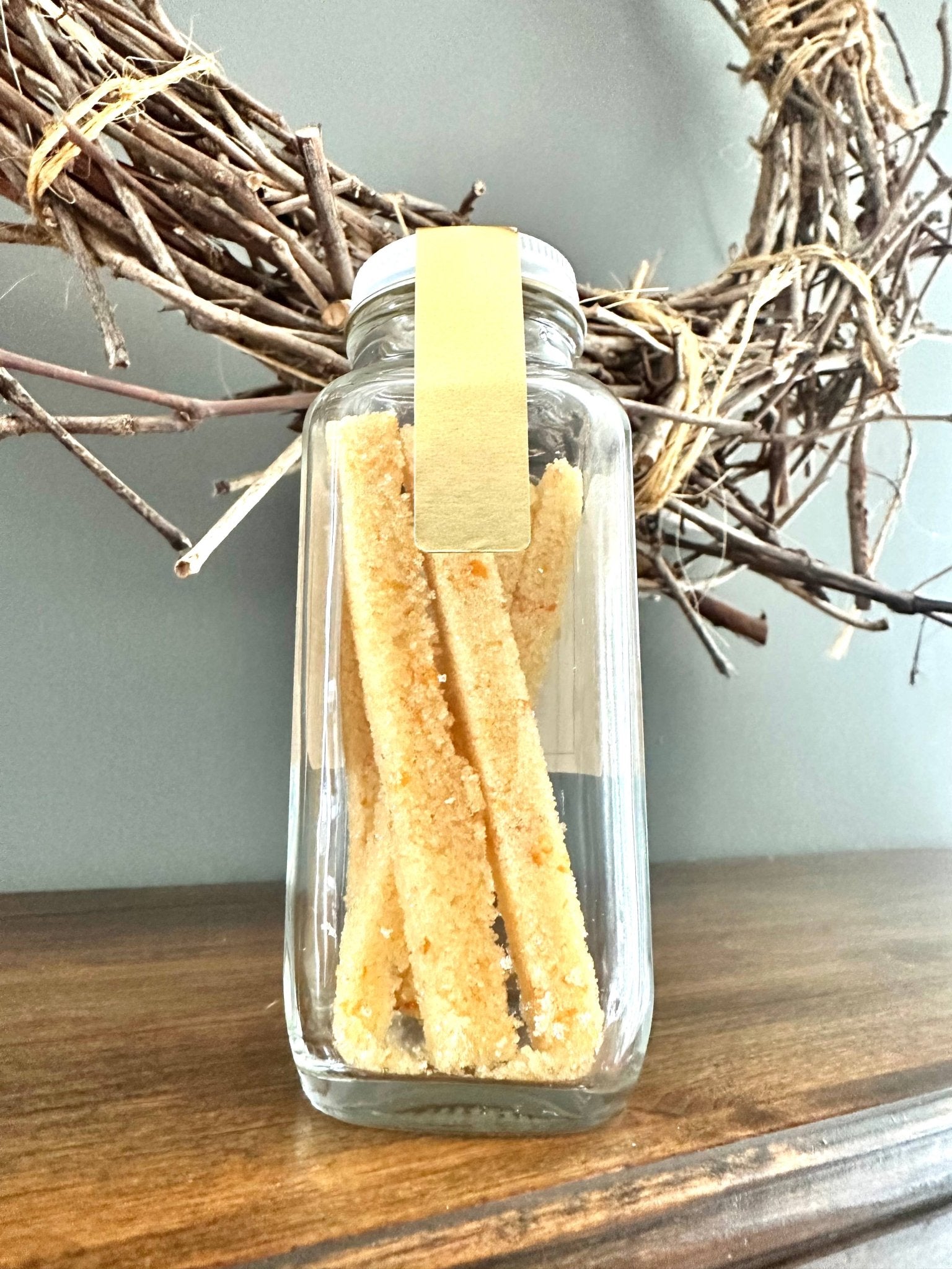 Clear glass jar filled with golden shortbread cookie sticks, with a white lid and yellow label, on a wooden surface.