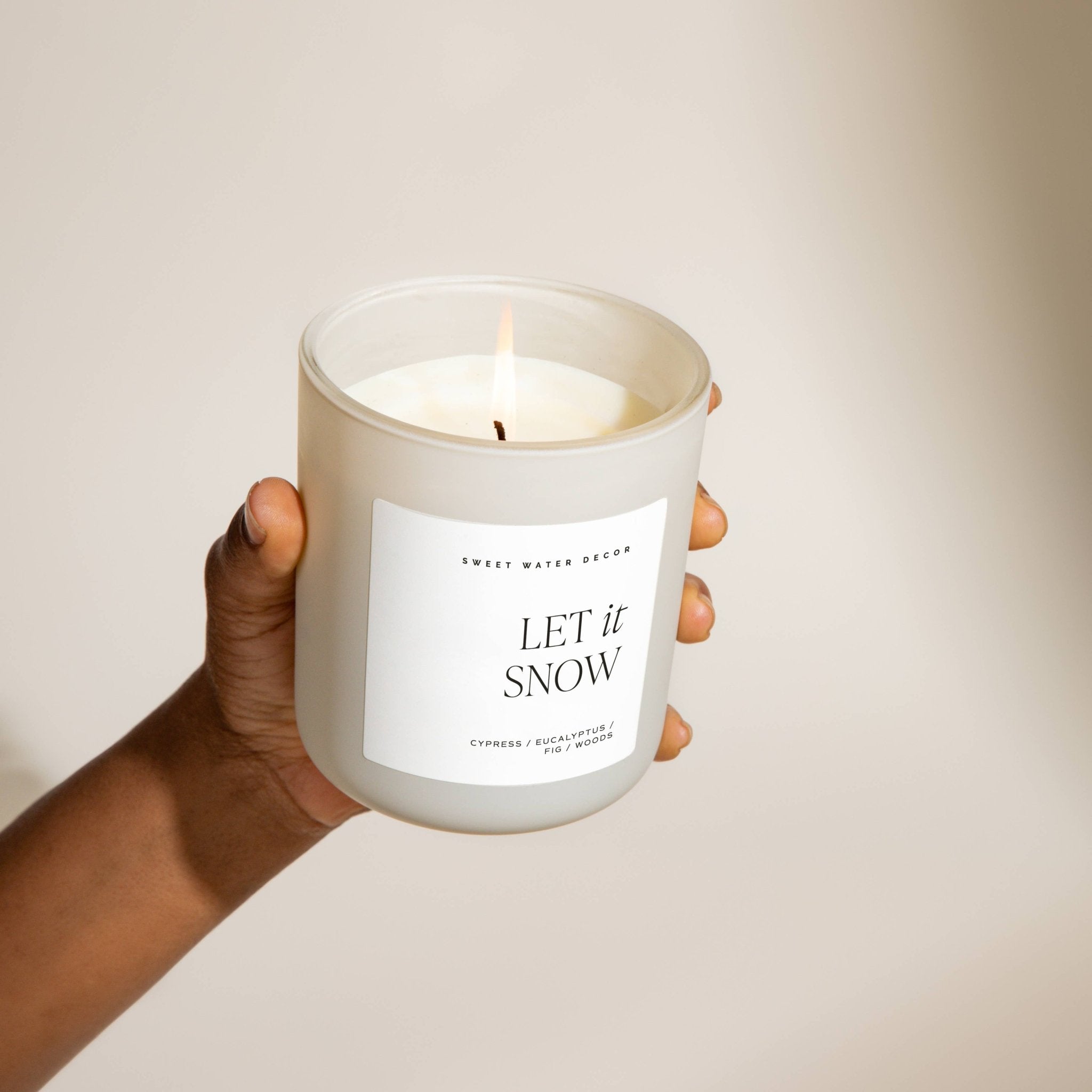 Candle labeled 'Let it Snow' held by a hand against a neutral background