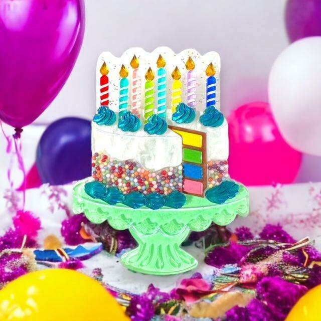 A colorful birthday cake decoration or toy cake on a green pedestal stand, surrounded by balloons and party confetti in a festive setting.