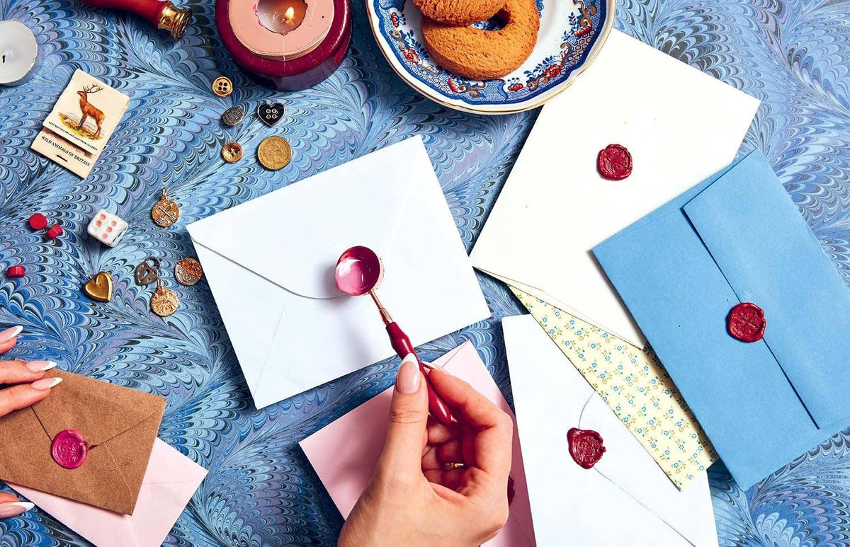 A styled flat-lay of wax seals and envelopes on a blue marbled surface, with a hand holding a red wax seal stamp. Visible items include white, blue, pink, and brown envelopes with red wax seals, scattered coins, a candle, and a small card.