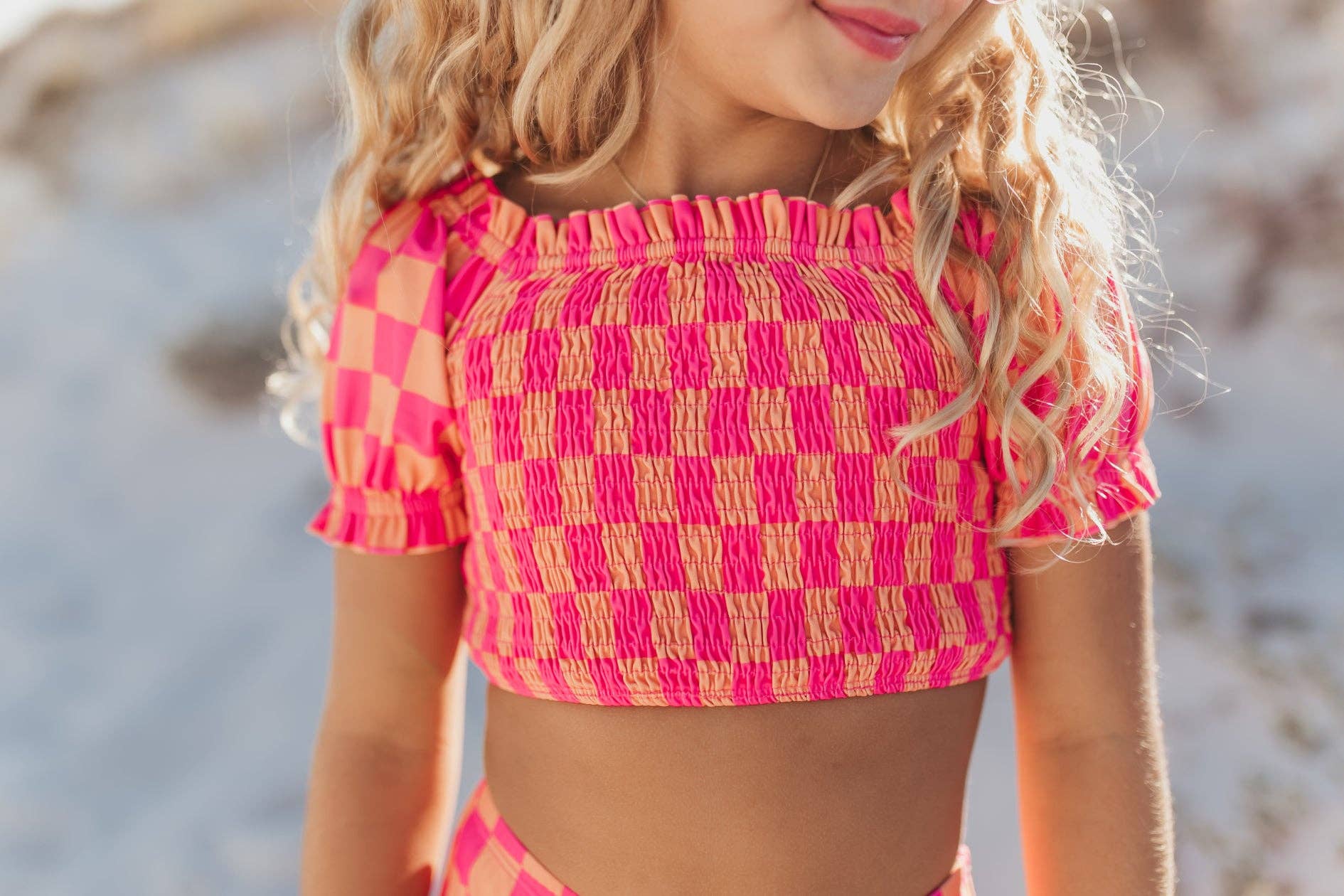 Girl in pink and orange checkerboard crop top with smocked bodice and ruffle sleeves.