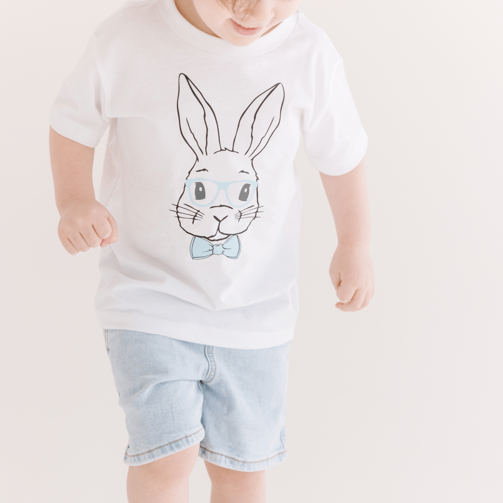 Toddler wearing a white t-shirt with a bunny graphic, blue glasses and bow tie, and light-wash denim shorts.