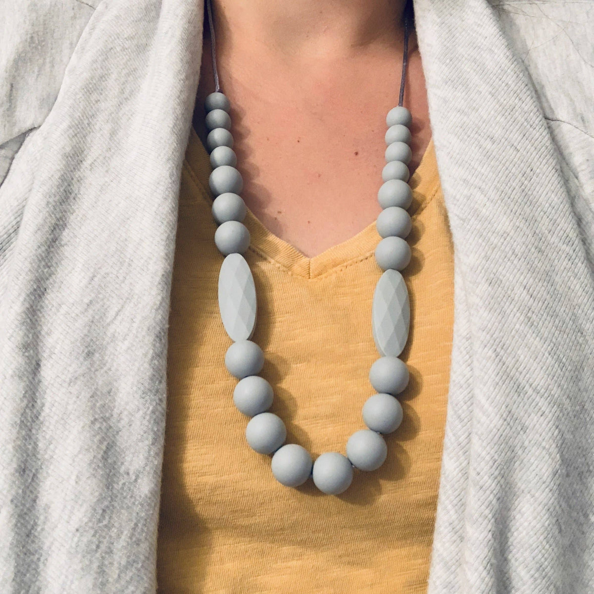 A close-up of a person wearing a light gray beaded necklace with two larger oval faceted beads, layered over a mustard yellow top and a light gray cardigan.