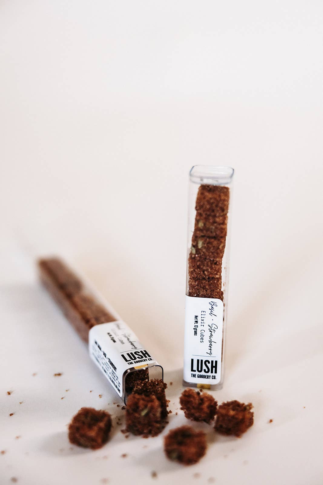 Two LUSH THE GOODERY CO. Basil Strawberry Elixir Cubes in clear tubes, one spilled, revealing brown granular cubes with green specks.