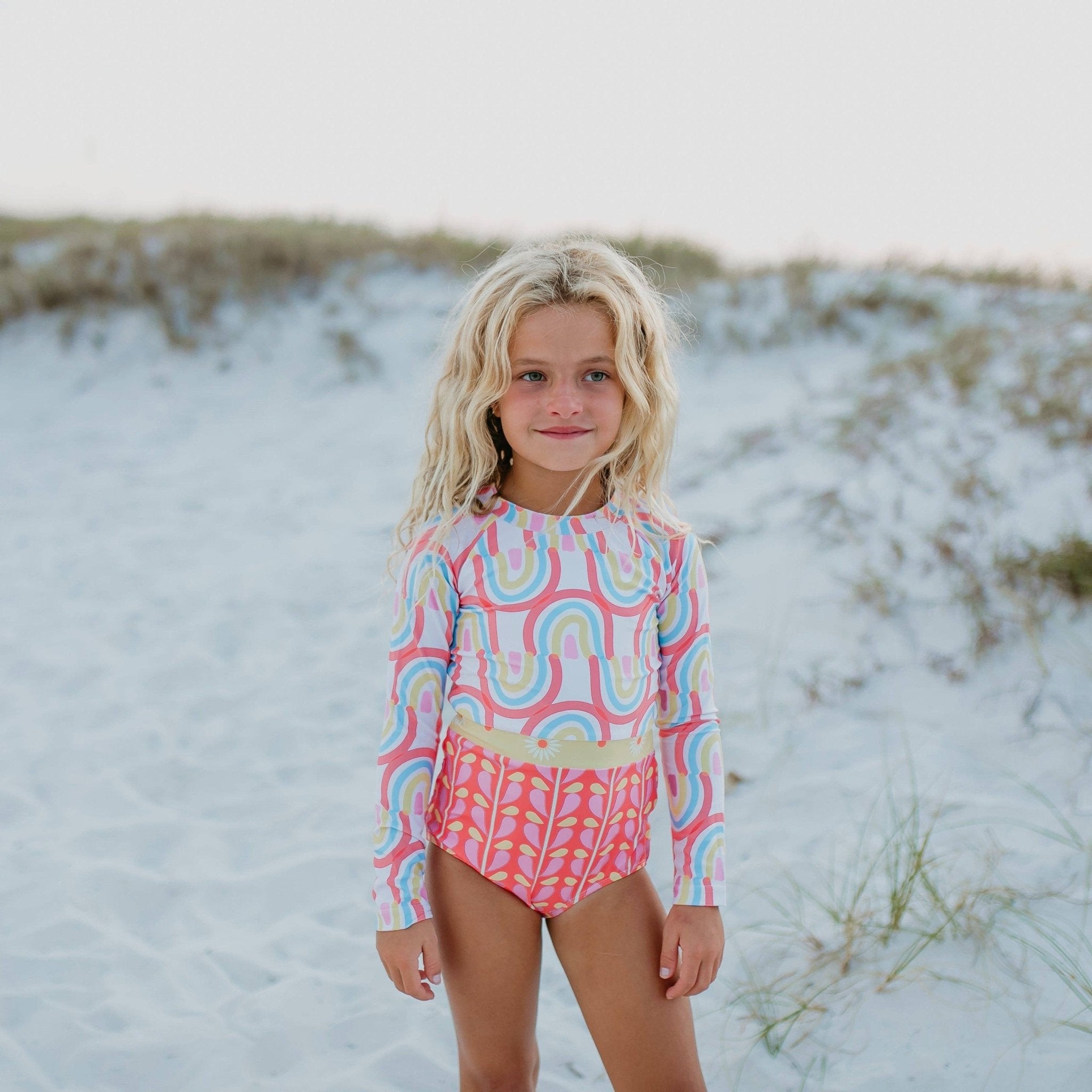 Girl in rainbow print long sleeve swimsuit with floral detail at the beach.
