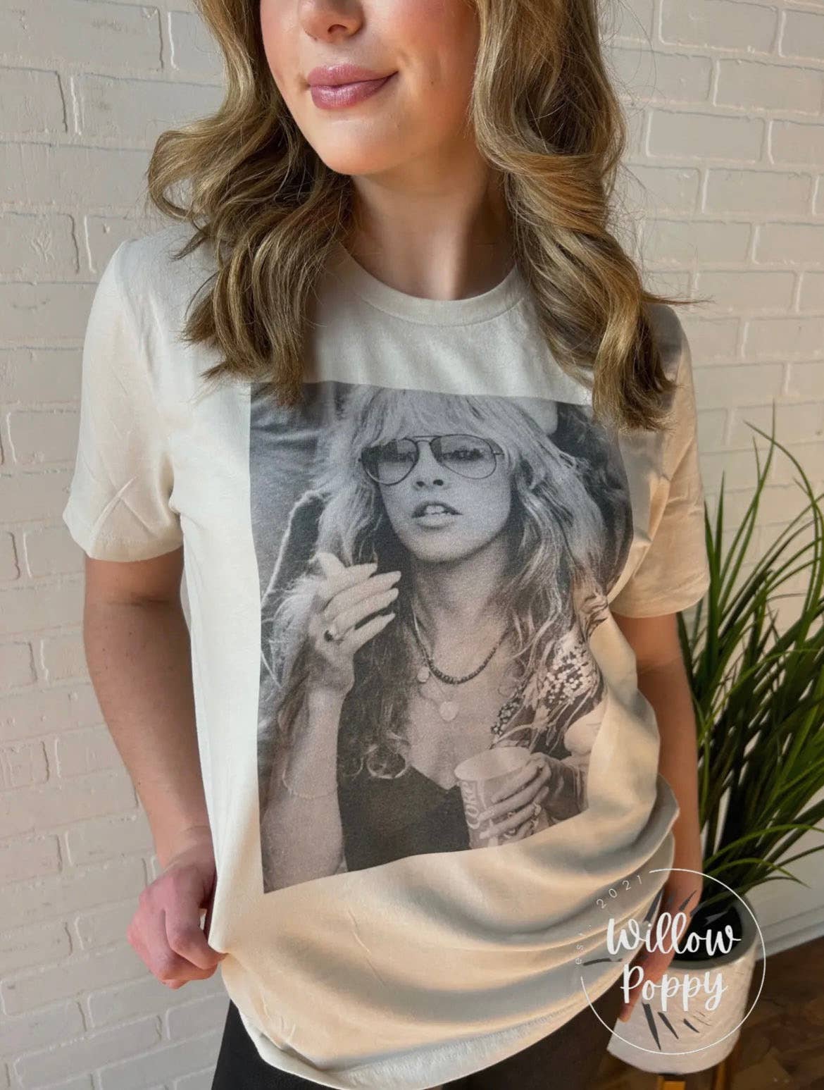Person wearing a t-shirt with a graphic design of Stevie Nicks face, standing against a white brick wall.