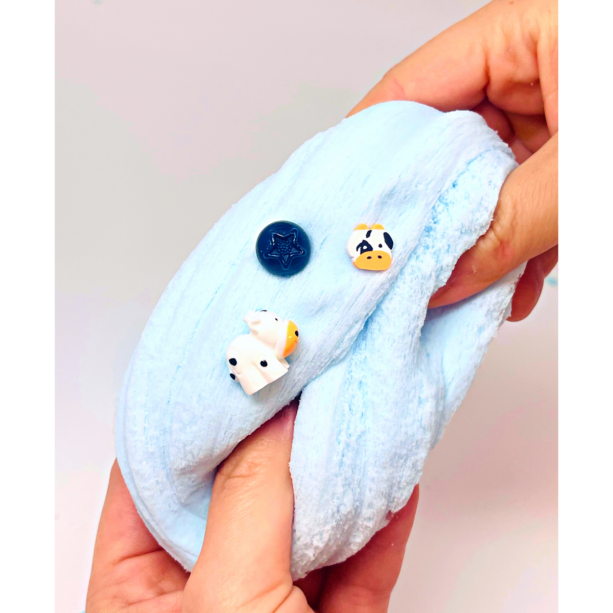 A close-up of a person's hands holding a light blue plush fabric item, possibly a small blanket, cushion, or pet toy, with three small decorative charms attached: a dark blue round charm, a white cow charm with black eyes, and a white duck charm with a yellow beak.
