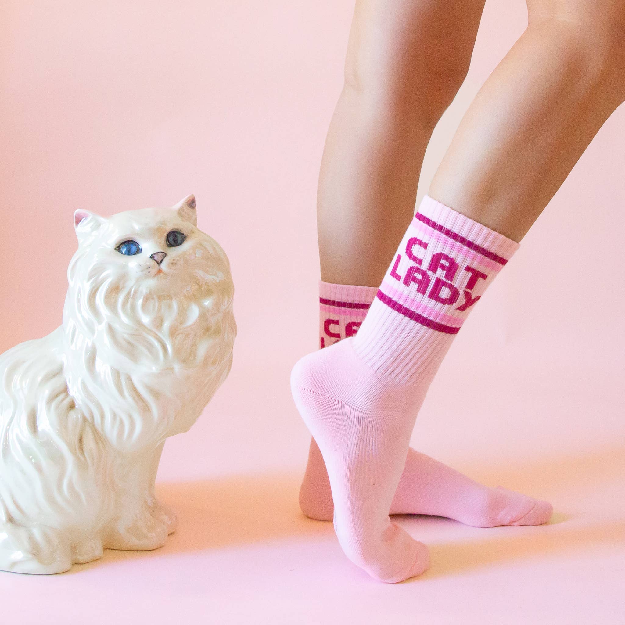 Pink 'Cat Lady' crew socks on a woman, with a white cat figurine on a pink background.