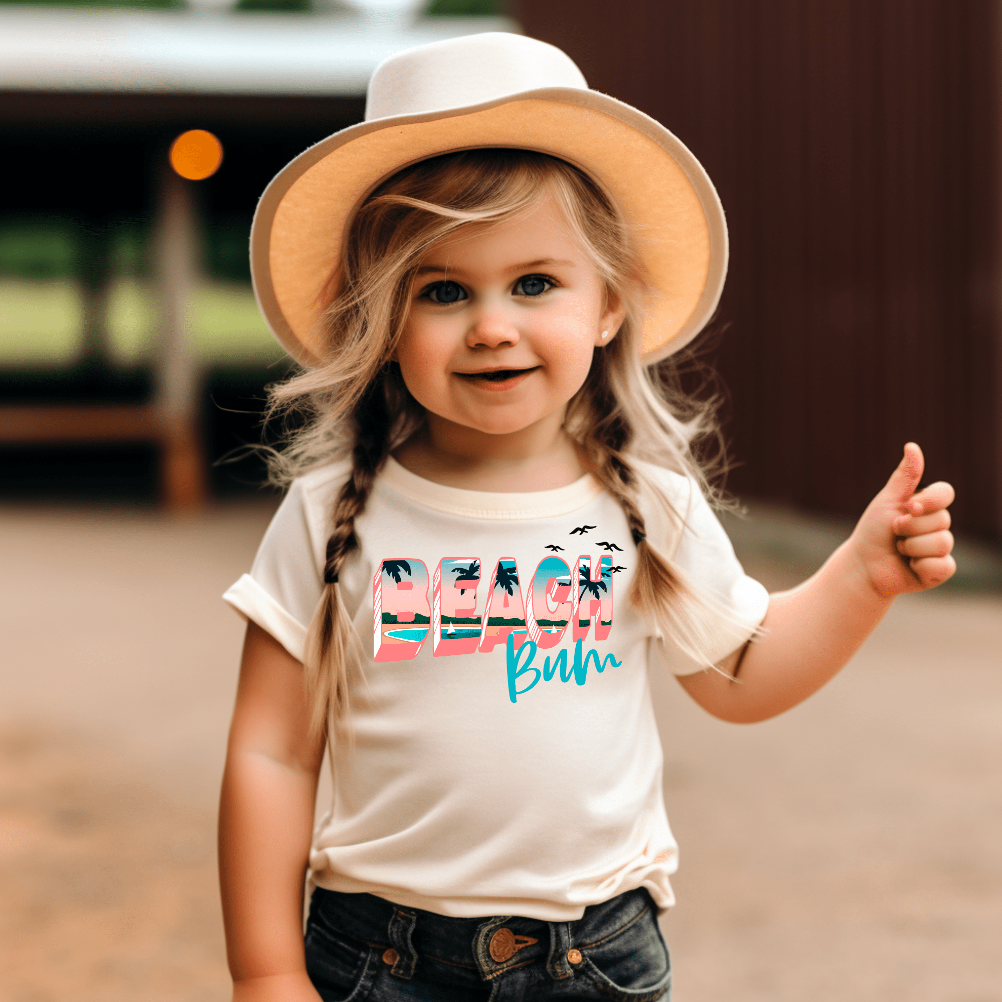 Toddler girl in a 'Beach Bum' graphic tee, straw hat, and jeans giving a thumbs up.