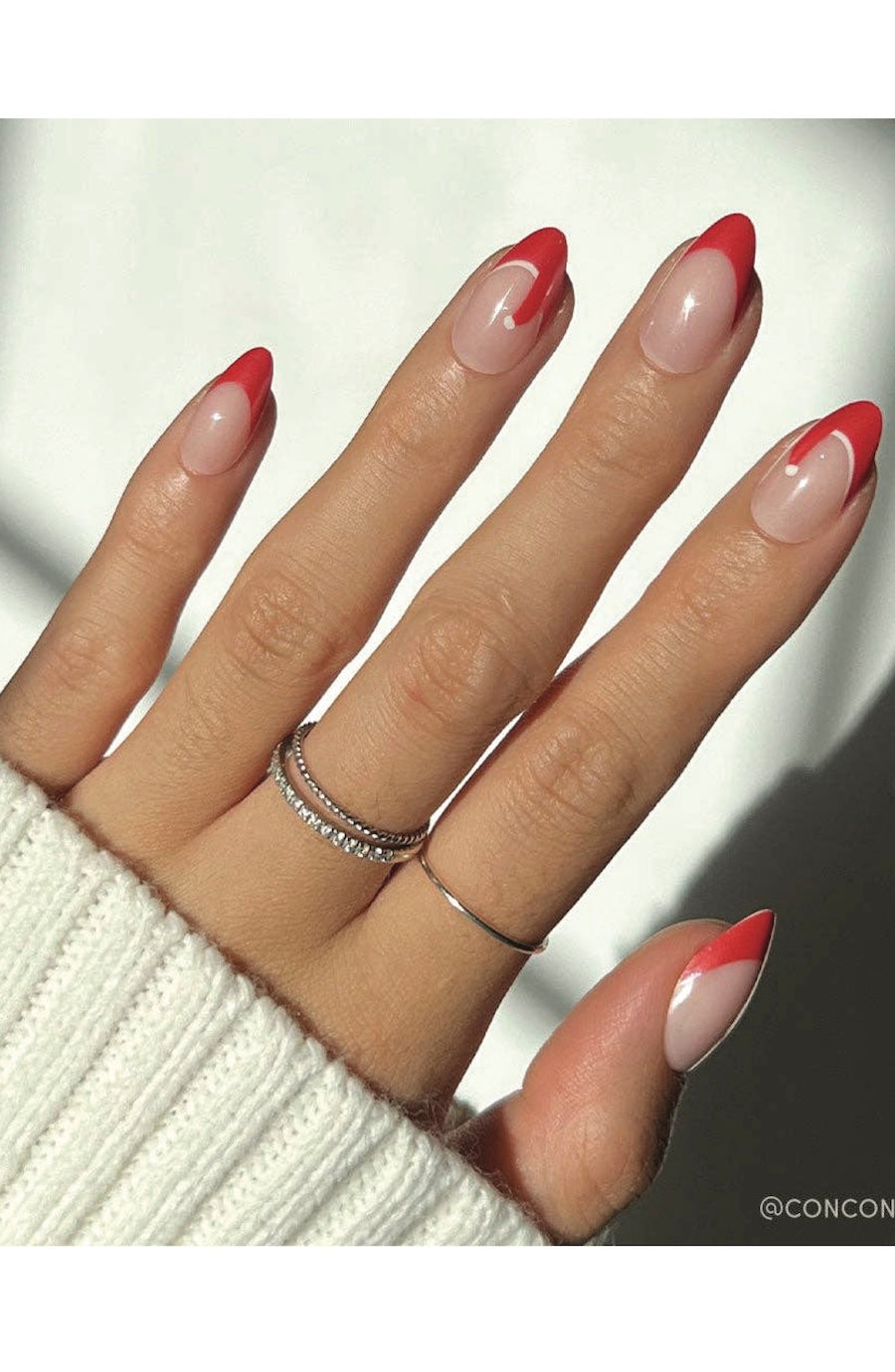 A close-up of a hand with long almond-shaped nails featuring a red and white French tip design, wearing two silver rings, including one with small clear stones.