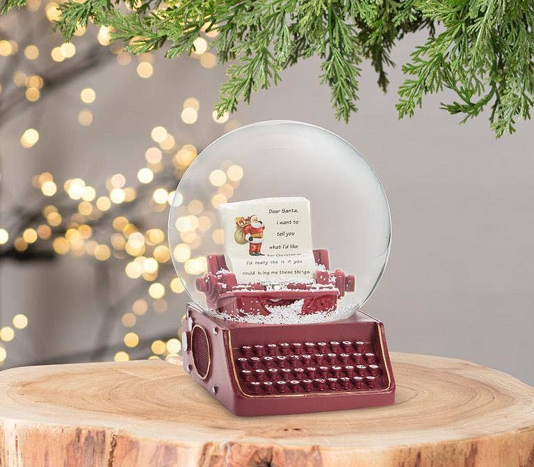 A Christmas-themed snow globe music box placed on a wooden slice, featuring a burgundy typewriter base, a clear glass dome with faux snow, and a handwritten letter inside. The background has warm bokeh lights and green pine branches.