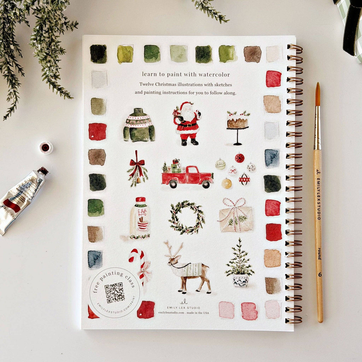 A spiral-bound watercolor painting book titled 'Learn to paint with watercolor' featuring Christmas illustrations, styled on a white surface with a paint tube, brush, and greenery.