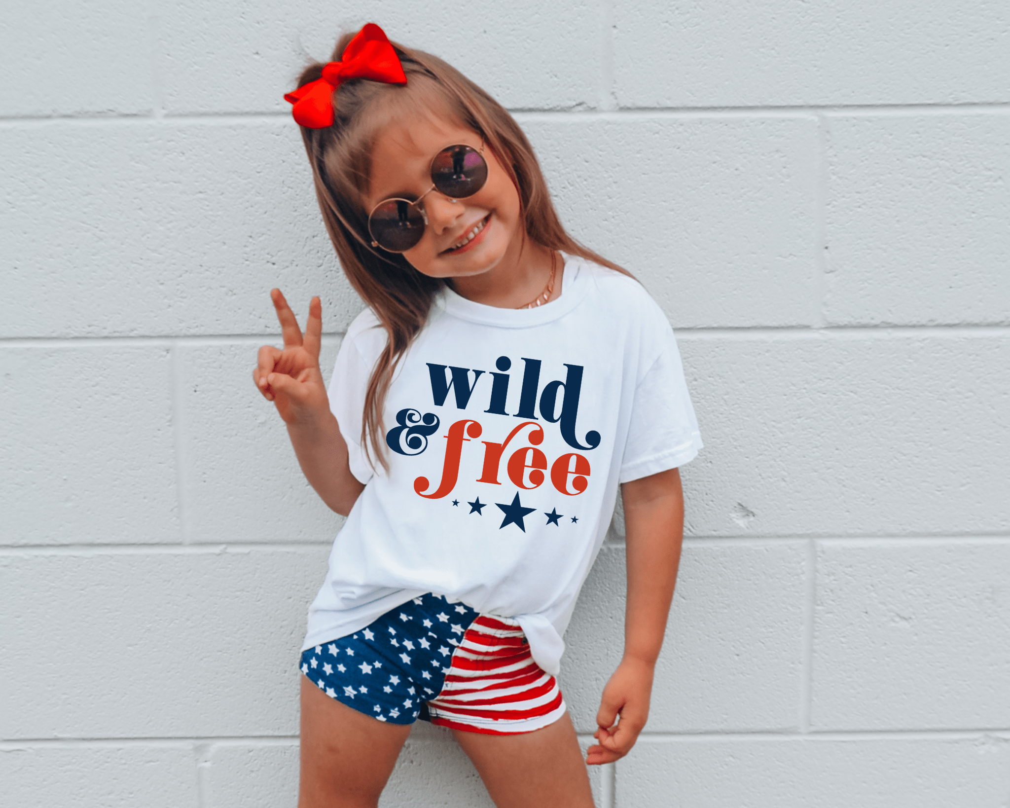 Girl in 'Wild & Free' 4th of July T-shirt and American flag shorts, red bow, sunglasses.