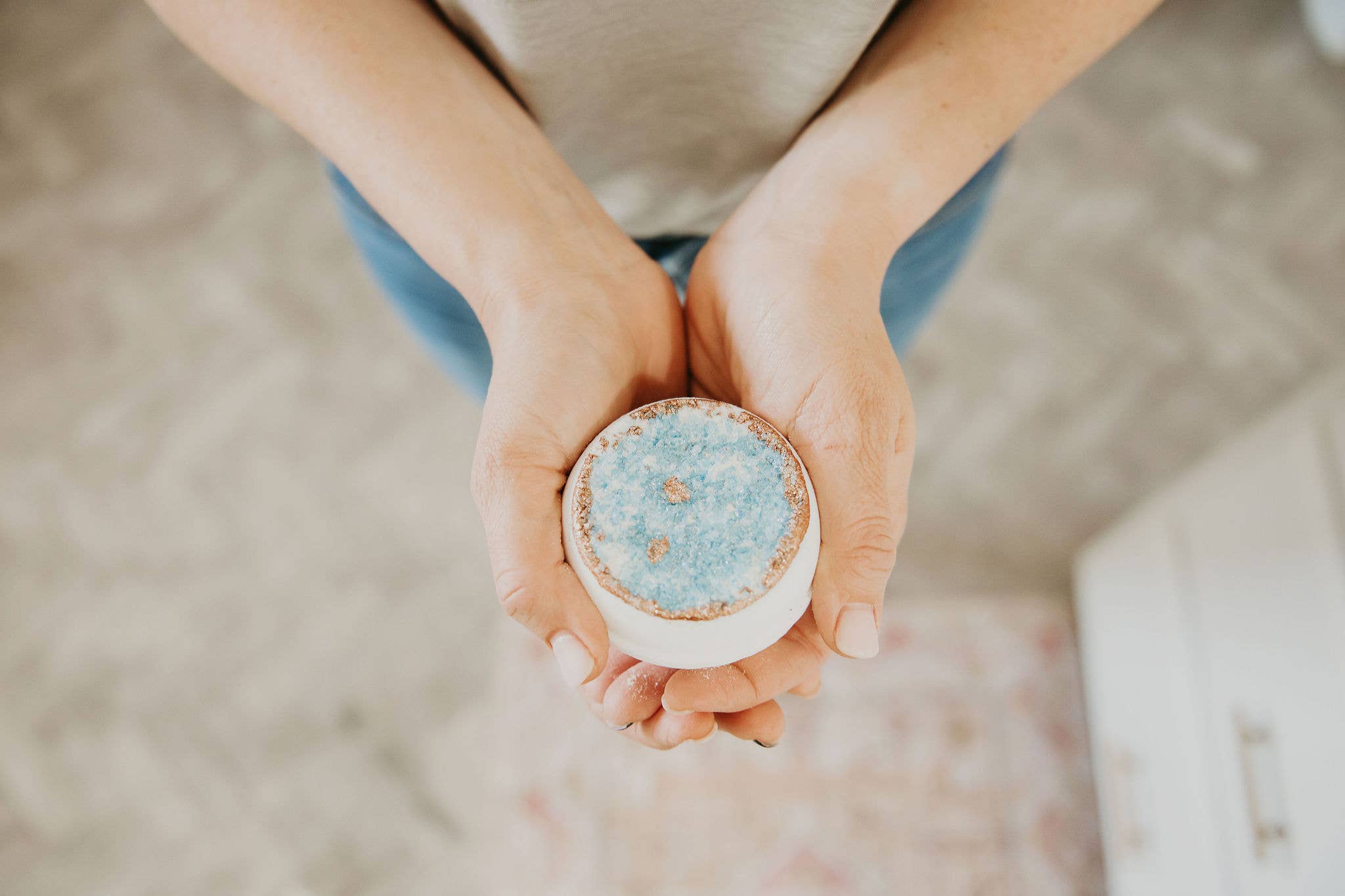 Hands holding a round, white bath bomb with a blue, textured top and gold flakes.