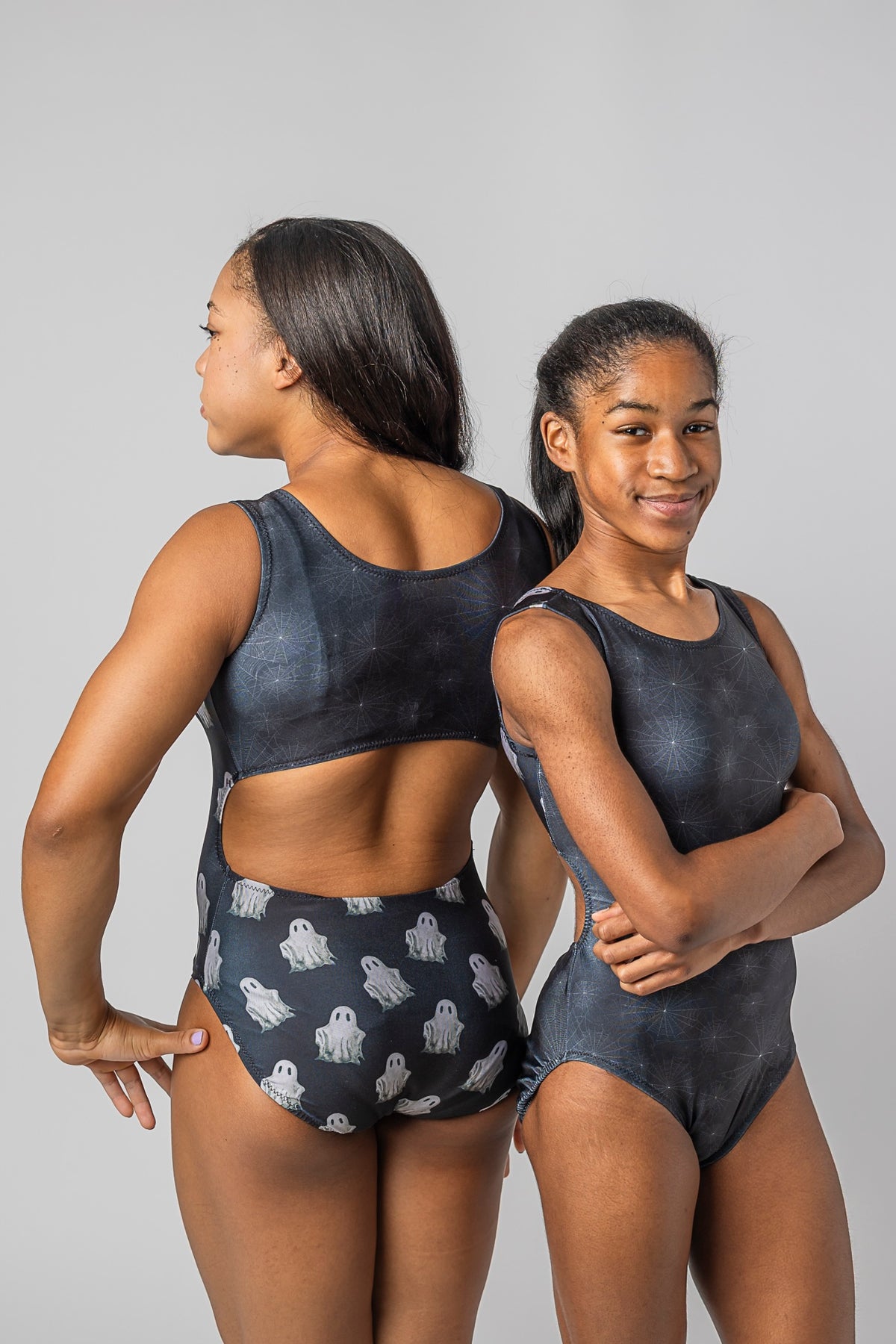 Two young gymnasts modeling matching dark gray and black leotards with white ghost patterns, posed against a solid light gray background.