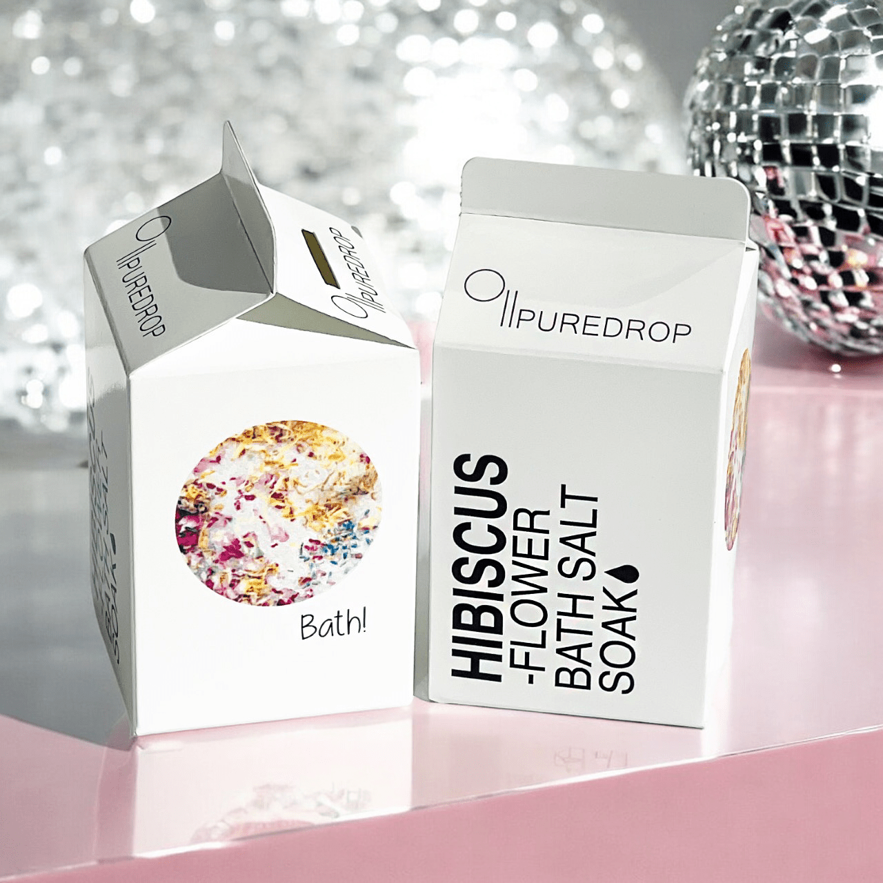 Two PUREDROP Hibiscus Flower Bath Salt Soak cartons on a pink surface, one showing dried petals.