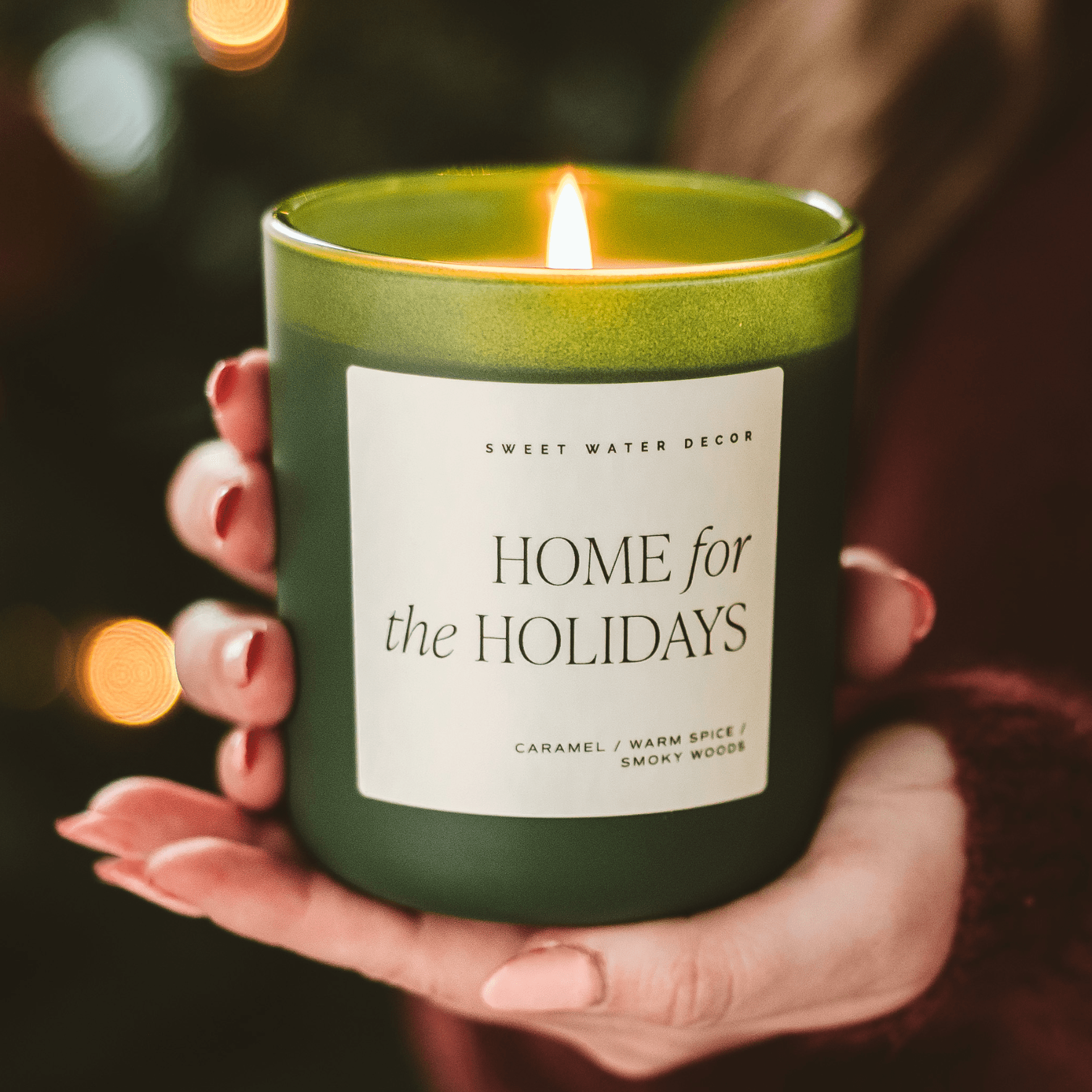 Green candle labeled 'Home for the Holidays' held in hands with a blurred background