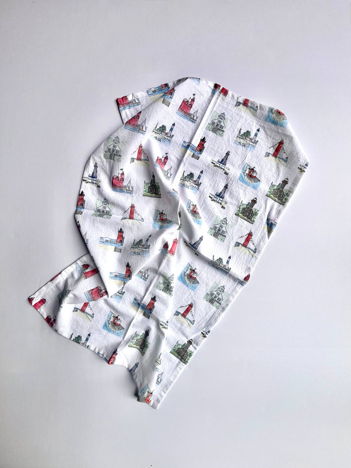 A white muslin baby blanket with a colorful winter nautical print of sailboats, lighthouses, and snowy trees, laid flat on a light gray background.