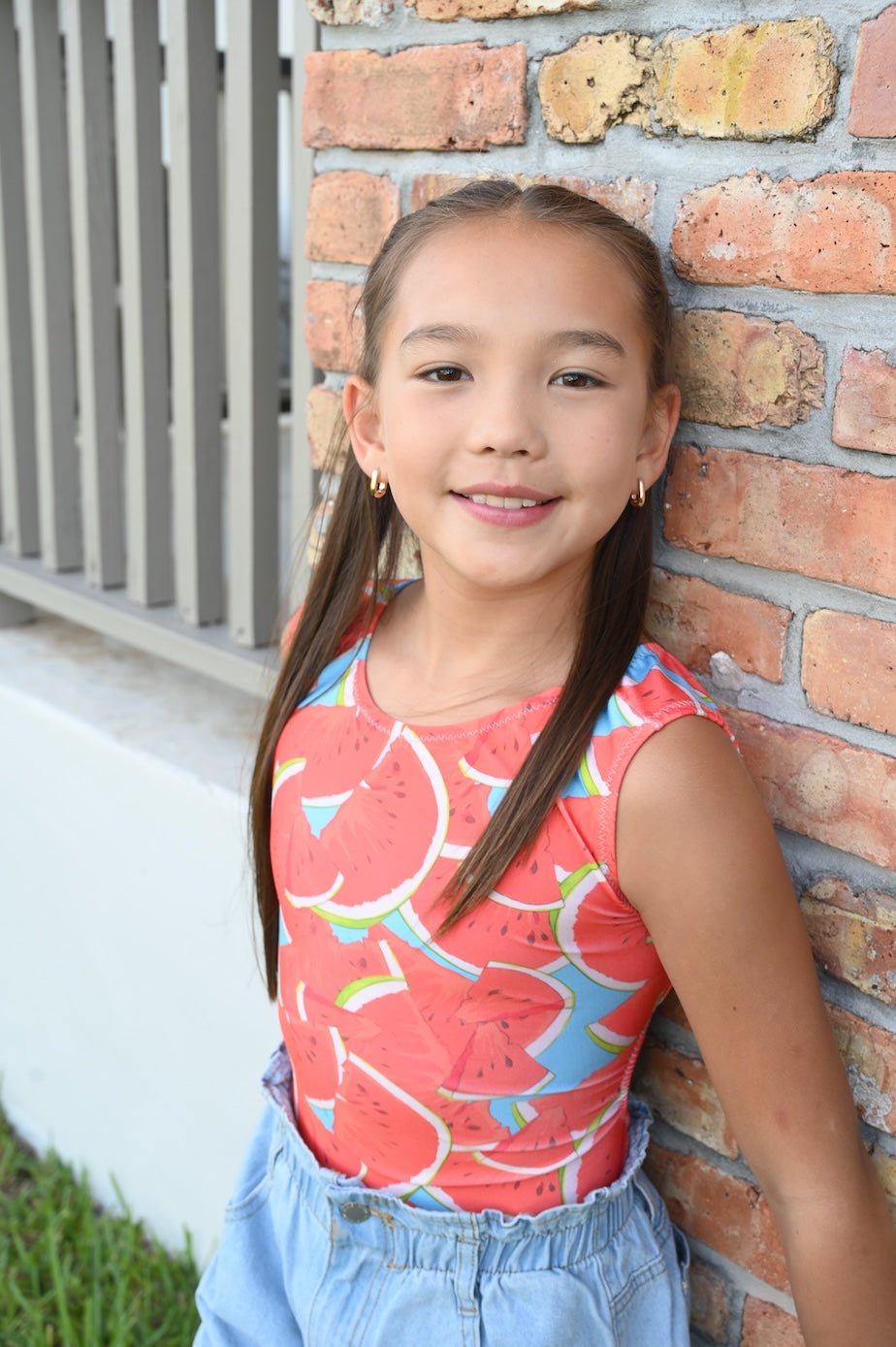 Young gymnast in a colorful watermelon gymnastics leotard leaning against a brick wall.