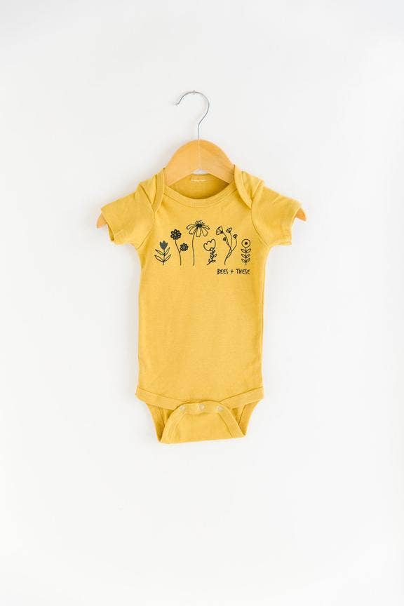 Mustard baby onesie with 'Bees + These' floral design on a hanger.