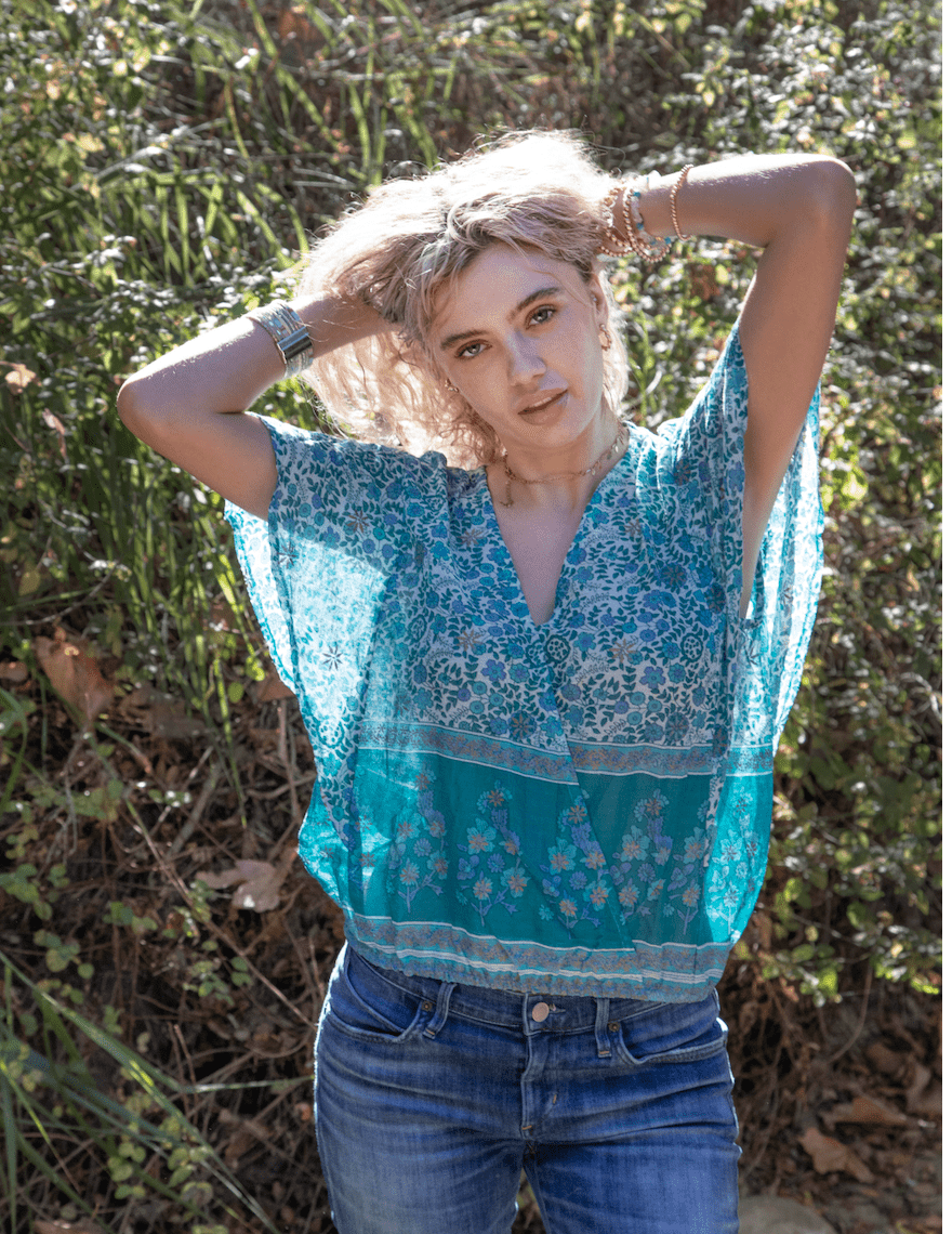 Woman wearing a blue patterned top and jeans standing outdoors with greenery in the background
