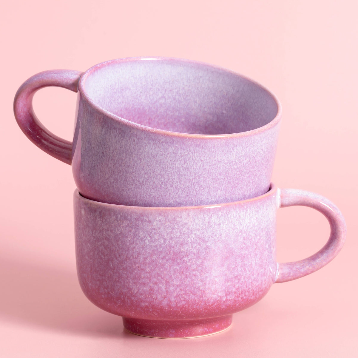 Two stacked ceramic mugs with a speckled lavender-pink glaze, set against a solid light pink background.