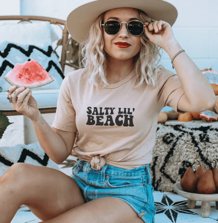 Peach t-shirt with 'Salty Lil' Beach' text. Casual summer style.