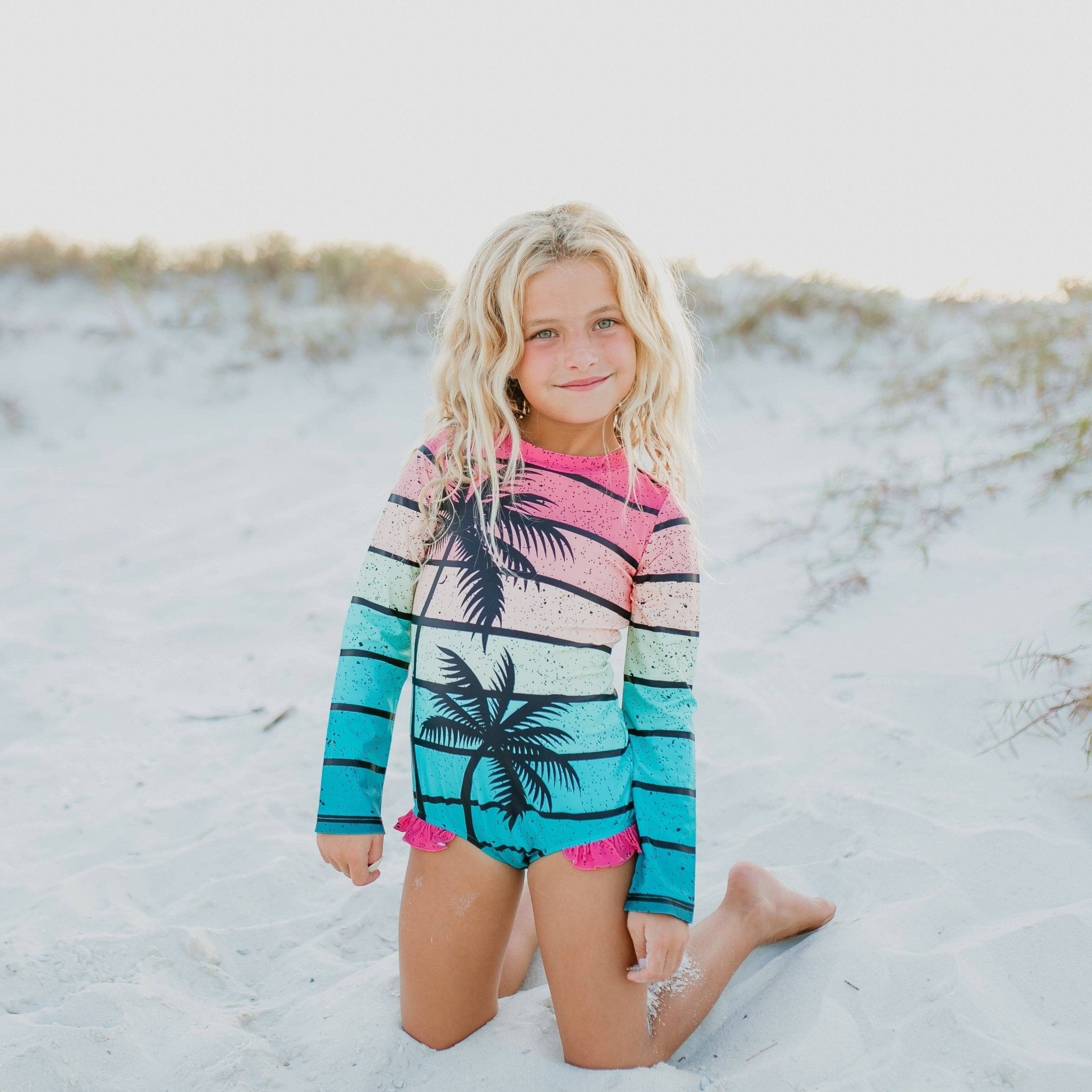Girl in palm tree rash guard swimsuit. Long-sleeve, striped surf suit in pink, blue, and green with palm tree design.