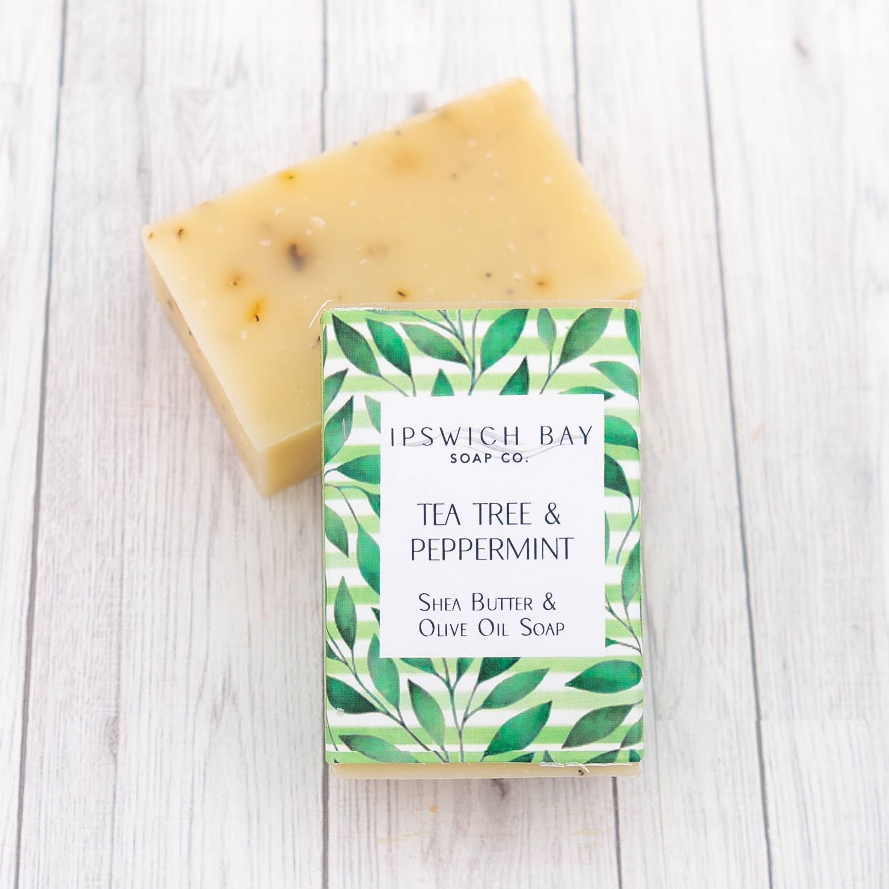 A bar of Tea Tree & Peppermint soap with a slice cut out, revealing the light yellow color of the soap. The packaging is green with a leaf pattern and the product name is clearly visible.