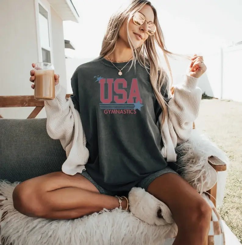 Woman wearing a 'USA Gymnastics' shirt, sitting outdoors holding a drink.