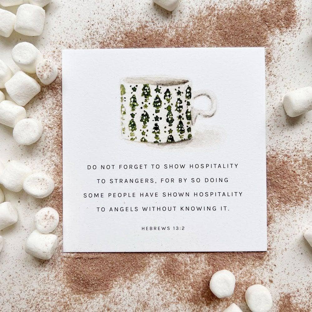 A square white card with a printed mug illustration and the text 'DO NOT FORGET TO SHOW HOSPITALITY TO STRANGERS, FOR BY SO DOING SOME PEOPLE HAVE SHOWN HOSPITALITY TO ANGELS WITHOUT KNOWING IT. HEBREWS 13:2' is photographed on a light surface with scattered marshmallows and cocoa powder.