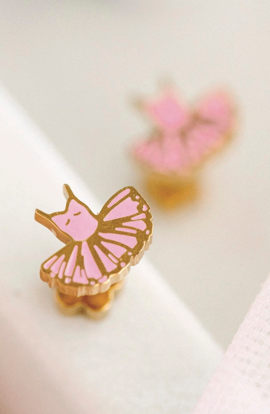 A close-up of a small ballerina-shaped stud earring with pink enamel and gold-tone metal, resting on a white surface with a blurred second earring in the background.