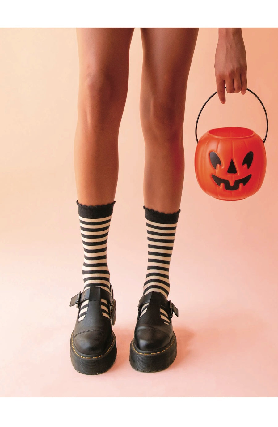 A person from the knees down wearing black-and-white striped crew socks and black Mary Jane platform shoes, holding a small orange jack-o'-lantern bucket against a soft peach background.