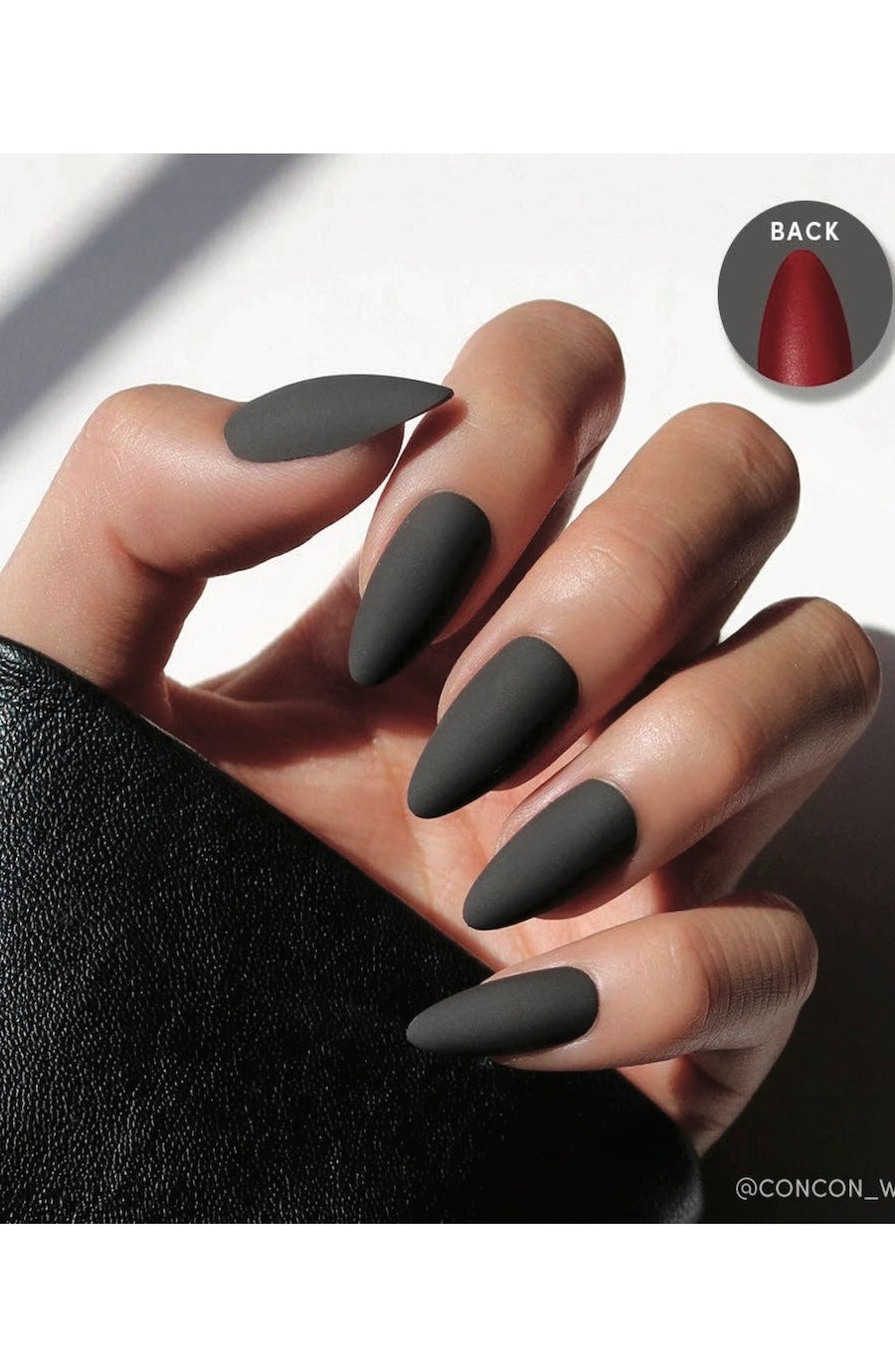 A hand with matte dark gray stiletto-shaped press-on nails, shown against a white background with a circular inset displaying the red backing of one nail.