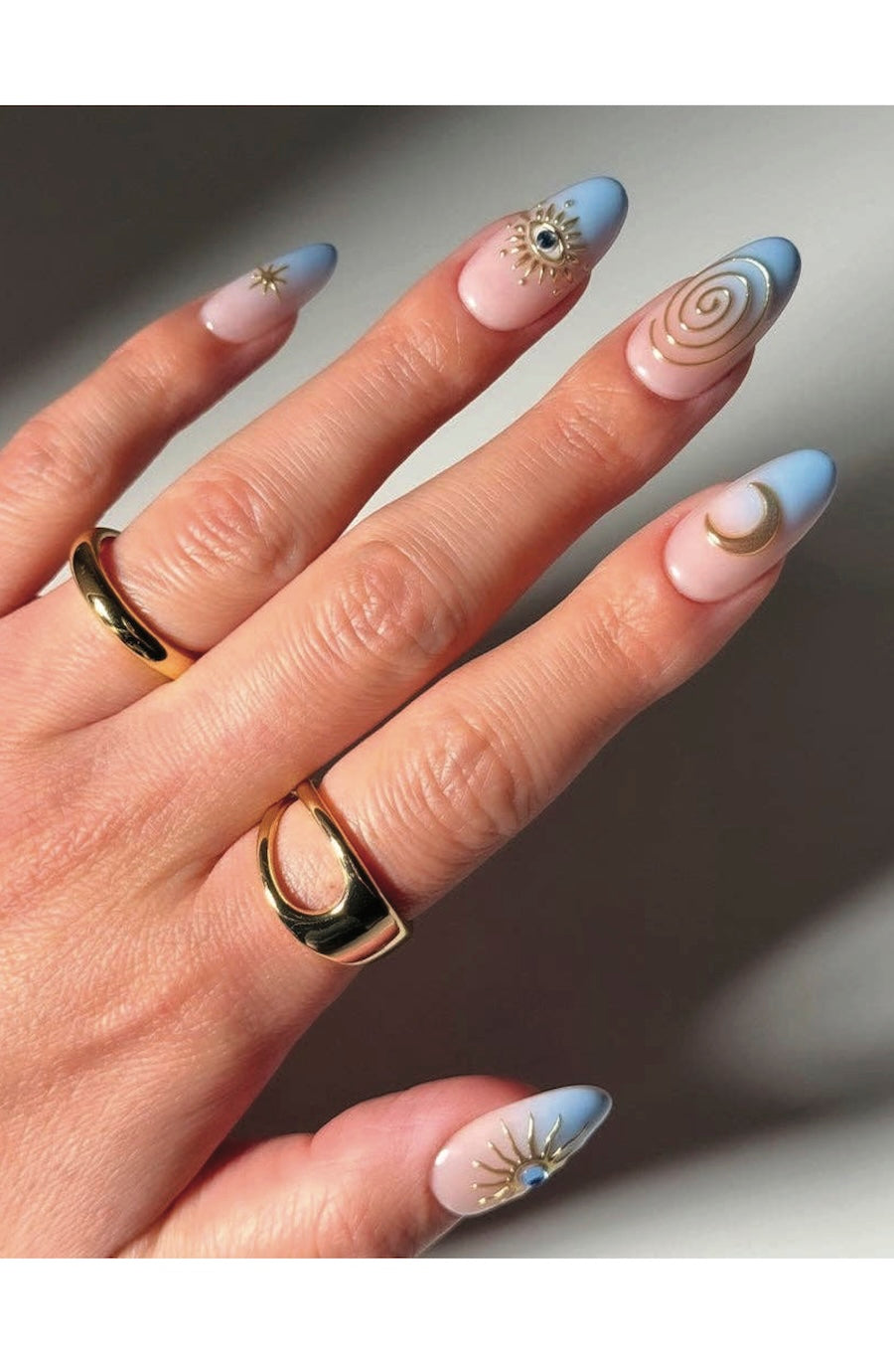 A close-up of a hand with manicured nails featuring a pastel blue and pink gradient, gold celestial designs, and two gold rings.