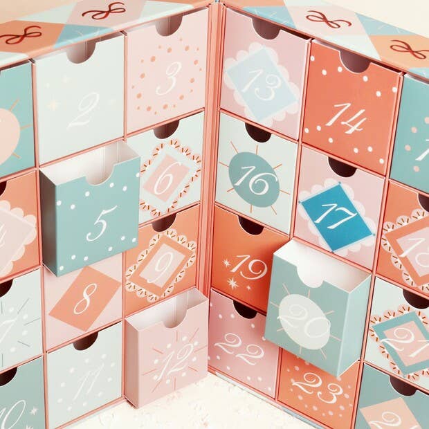 A pastel advent calendar made of many small numbered drawers with pull tabs, shown in a clean studio-style close-up. The drawers are printed with soft pink, peach, mint, and light blue patterns and white numbers.