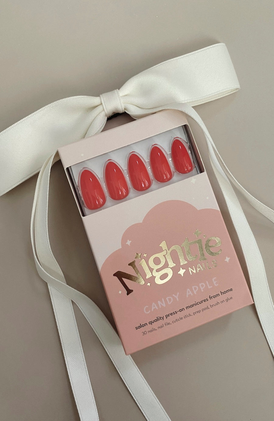 A boxed set of false press-on nails tied with a white satin ribbon. The nails are a glossy coral-red shade, and the box is pale pink with metallic gold lettering that reads 'Nightie Nails' and 'Candy Apple'.