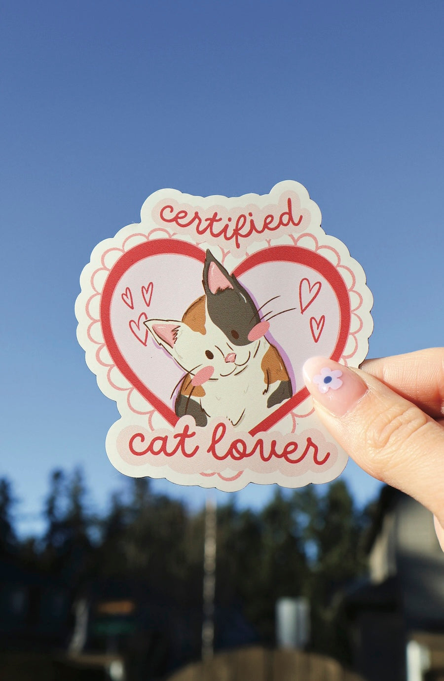 A hand holds a scalloped die-cut sticker featuring a cartoon calico cat inside a red heart, with the words 'certified cat lover' in red script, against a blurred outdoor background with blue sky.