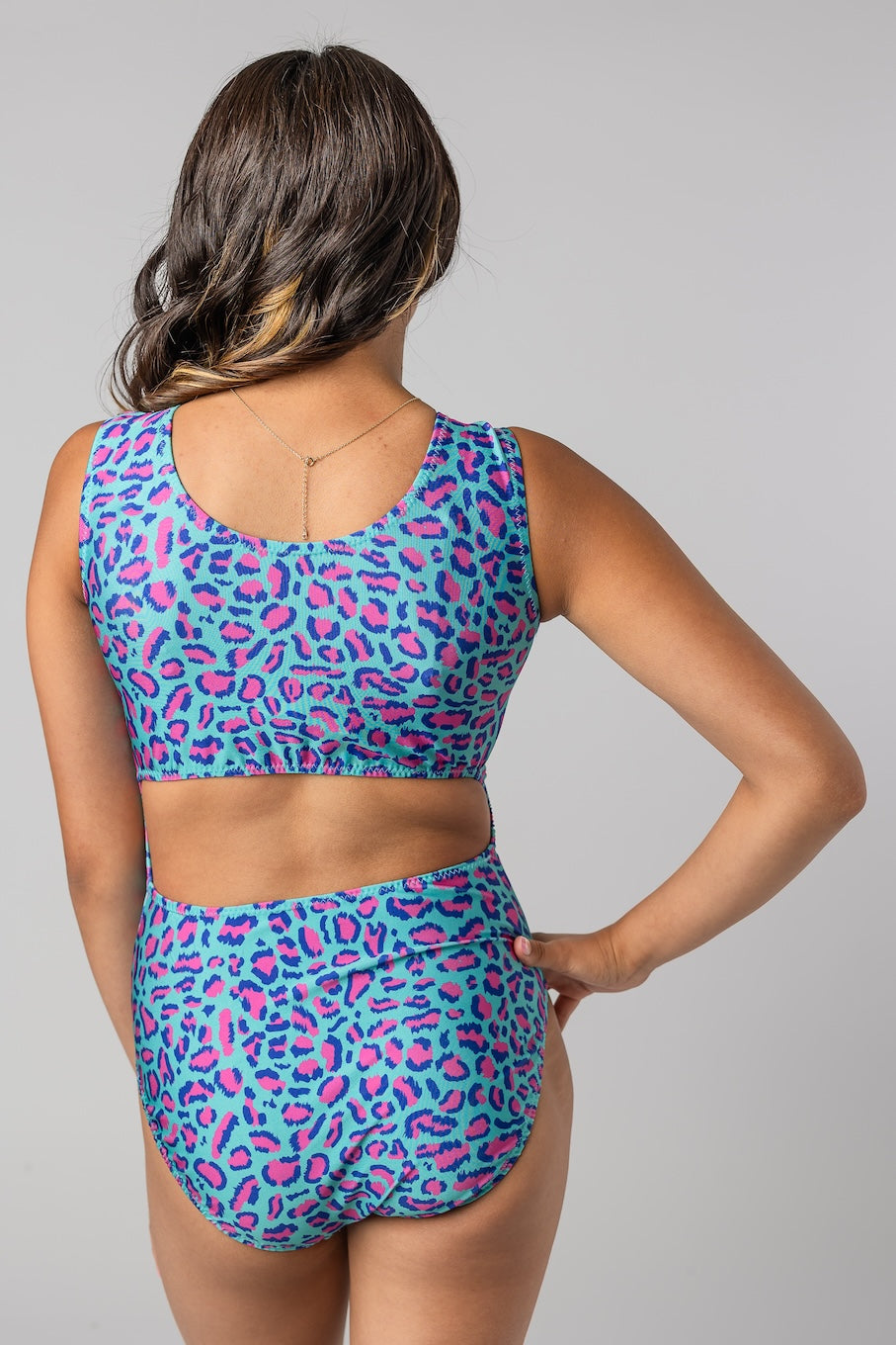 A woman shown from the back wearing an open-back leotard with bright blue, pink, and purple leopard print pattern against a solid gray studio background.