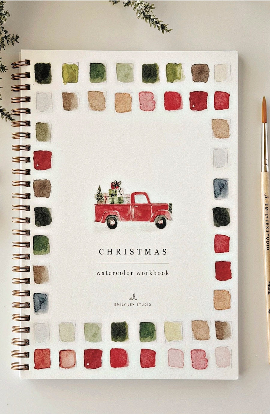 A spiral-bound watercolor workbook titled "Christmas watercolor workbook" by Emily Lee Studio, featuring a red truck illustration and a border of watercolor color swatches, shown on a light surface with a paintbrush and greenery.