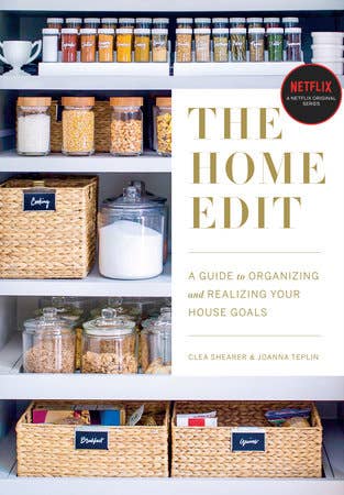 Organized pantry featuring 'The Home Edit' book, labeled wicker baskets, and clear jars filled with spices and grains.