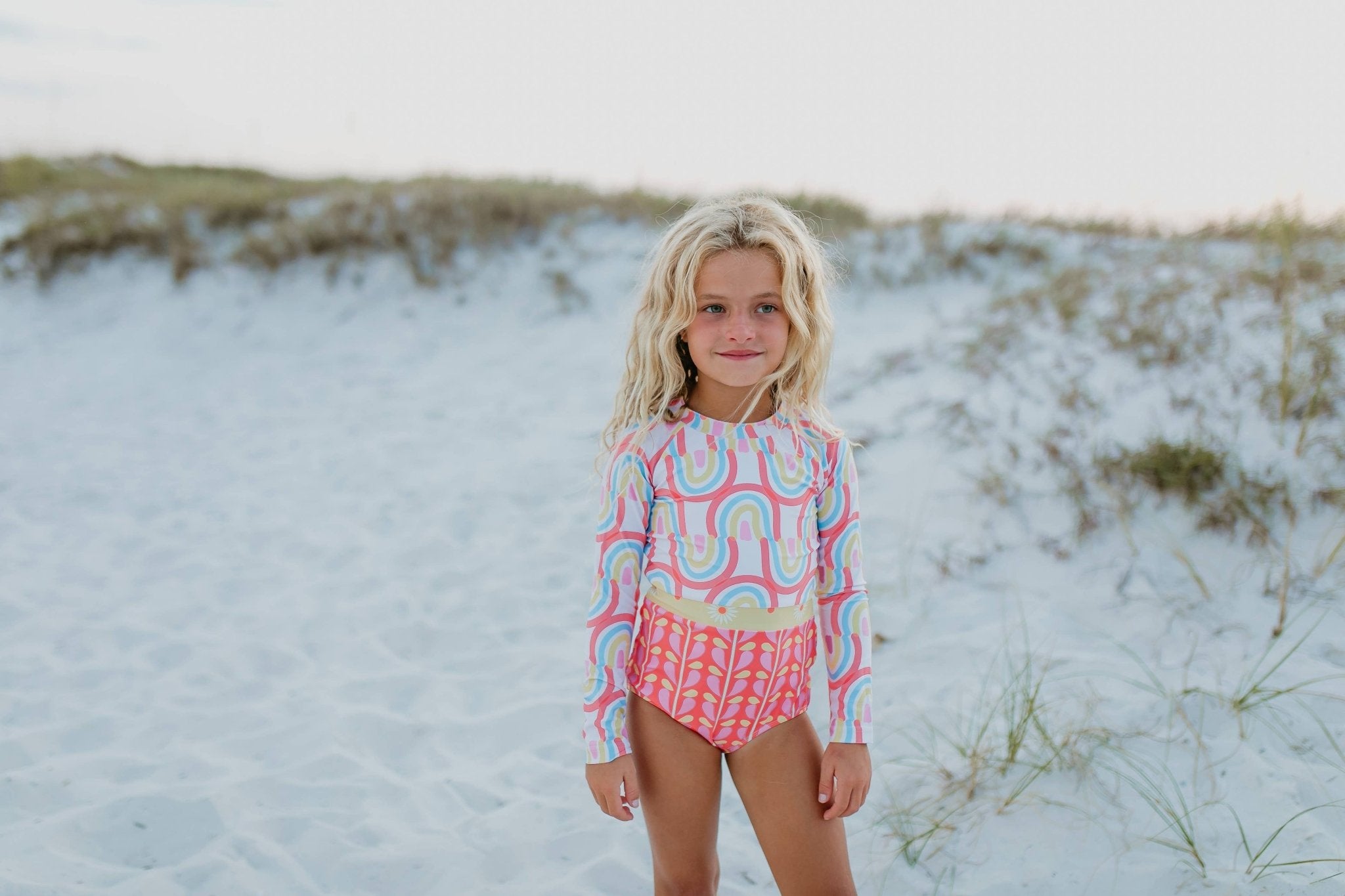 Girl in rainbow and floral long-sleeve swimsuit on beach.