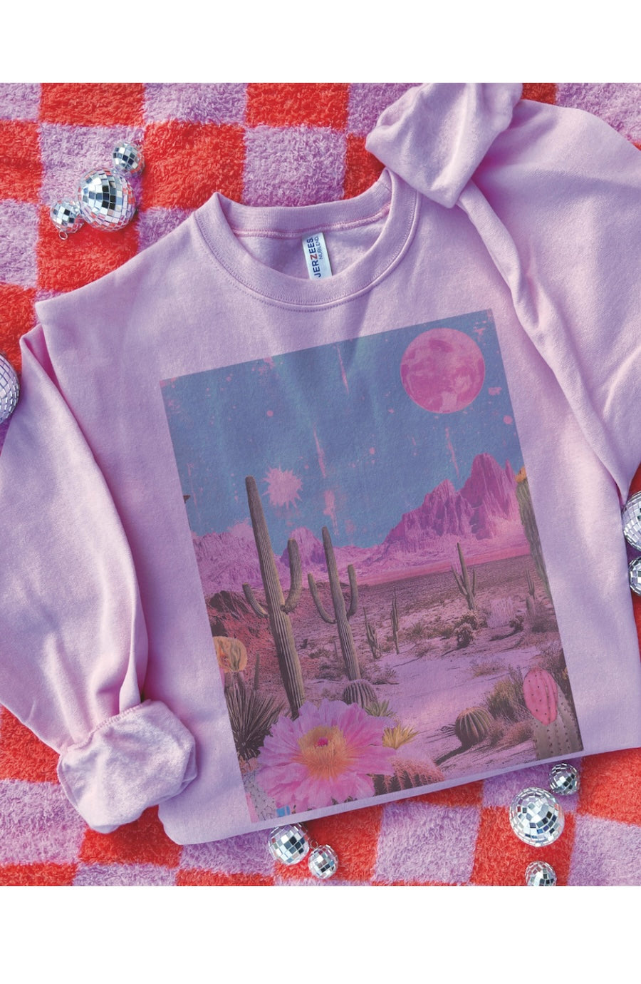 A lavender long-sleeve sweatshirt with a colorful retro desert landscape graphic, laid flat on a red and pink checkered blanket with silver disco balls.